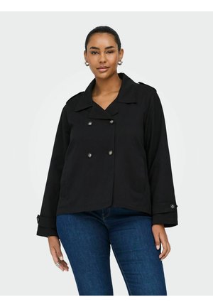 Kobieta ubrana w czarną dwurzędową marynarkę z zapiętymi mankietami i ciemnoniebieskie dżinsy, stojąca na jednolitym jasnym tle.