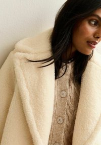 Cappotto oversized di colore crema, in peluche, con ampio colletto, indossato sopra un maglione lavorato a trecce di colore marrone chiaro, con quattro bottoni rotondi.