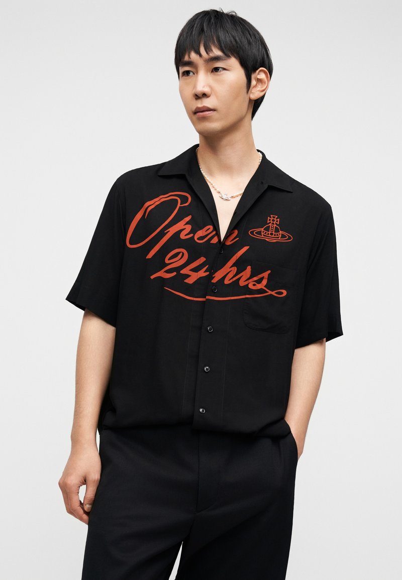 Chemise noire à manches courtes avec un imprimé graphique rouge indiquant "Ouvert 24h" et une petite poche poitrine, fabriquée dans un tissu lisse.