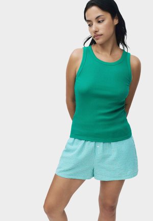 Frau steht mit den Armen hinter dem Rücken, trägt ein ärmelloses grünes, geripptes Oberteil und hellblaue, karierte Shorts vor einem schlichten Hintergrund.