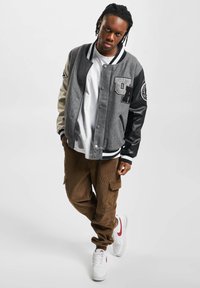 Grå og svart varsityjakke med skinnarmer, fremtredende bokstaver, kombinert med brune kordfløyels cargo bukser og hvite joggesko med røde detaljer.