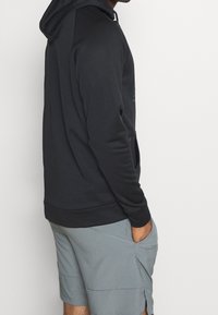 Felpa nera con cappuccio dalla texture liscia, tasca a marsupio e polsini a costine, abbinata a pantaloni corti grigi chiari.