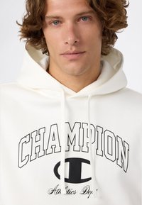 Giovane uomo con capelli ricci che indossa una felpa bianca Champion con un grande logo nero e cordini, su uno sfondo semplice.
