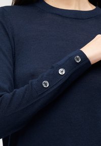 Ναυτικό μπλε πλεκτό πουλόβερ με στρογγυλή λαιμόκοψη και τρία ασημένια κουμπιά στη μανσέτα, με απαλή υφή και εφαρμοστά μανίκια.