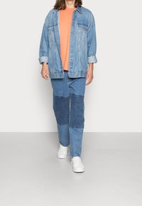 Osoba v světle modré džínové bundě přes oranžové tričko, s džínovými kalhotami patchwork a bílými teniskami, stojící před jednobarevným pozadím.