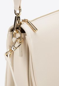 Bílá kožená taška se zlatými detaily, obsahující zip a karabinu. Hladký povrch s minimalistickým designem a nenápadným brandingem.