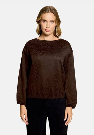 Femme aux cheveux blonds portant un chemisier marron foncé texturé à manches longues et un pantalon noir, face à un fond uni.