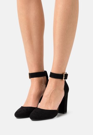 Chaussures à talons en daim noir avec un bout pointu, une bride à la cheville et un talon carré épais. Dotées d'une boucle dorée décorative sur la bride.