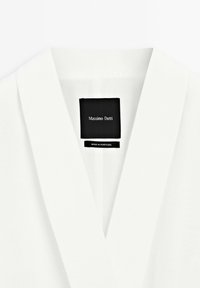 Vit blus med sjalkrage. Har en svart etikett med texten "Massimo Dutti" och "Made in Portugal". Slät, lättviktsmaterial.