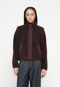 Fleecejacke in dunkelburgunderrot mit hohem Kragen, zwei Seitentaschen und einer glatten mittleren Blende. Strukturierter Stoff sorgt für Wärme und Komfort.