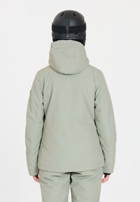 Hellgrüne isolierte Jacke mit Kapuze, matte Oberfläche und nahtlosem Design. Getragen mit einem schwarzen Helm, der einen Verschluss auf der Rückseite hat.
