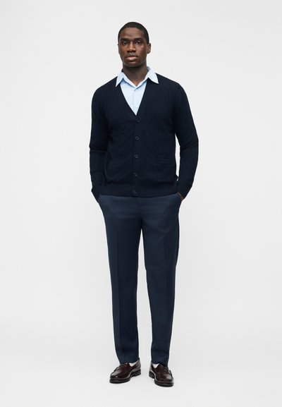 Άνδρας που φοράει ανοιχτό μπλε πουκάμισο, σκουρό μπλε ζακέτα με κουμπιά, ταιριαστό παντελόνι και καφέ δερμάτινα παπούτσια, στέκεται με τα χέρια στις τσέπες.