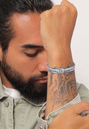 Silberner geflochtener Armreif mit einem Verschluss, getragen an einem Handgelenk, das komplexe Tätowierungen zeigt. Der Hintergrund ist neutral und hebt das Design des Armbands hervor.