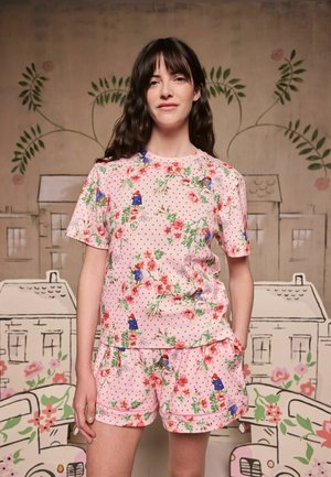 Femme aux longs cheveux foncés portant une chemise rose à manches courtes à motifs floraux et à pois ainsi qu'un short assorti, debout devant un fond illustré décoratif.