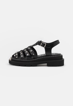 Schwarze Sandale mit dicker Sohle, silbernen Nieten auf geflochtenen Riemen, Schnallenverschluss am Knöchel und kräftigem Profil, gezeigt vor schlichtem weißen Hintergrund.