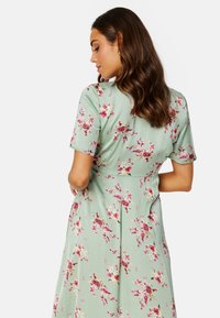 Kwiatowa sukienka w jasnozielonym materiale z krótkimi rękawami, ozdobiona różowymi i fioletowymi wzorami kwiatowymi, dopasowanym stanem i luźną spódnicą.