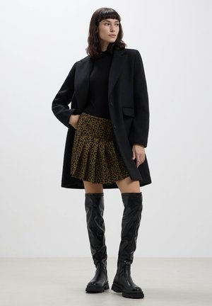Cappotto lungo nero su un maglione aderente nero, abbinato a una gonna a pieghe in stampa leopardata marrone e nera. Stivali lucidi neri fino al ginocchio.