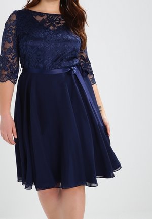 Granatowa sukienka z koronkowym gorsetem i rękawami, satynową wstążką w talii zawiązaną z boku oraz zwiewną spódnicą sięgającą kolan na osobie.