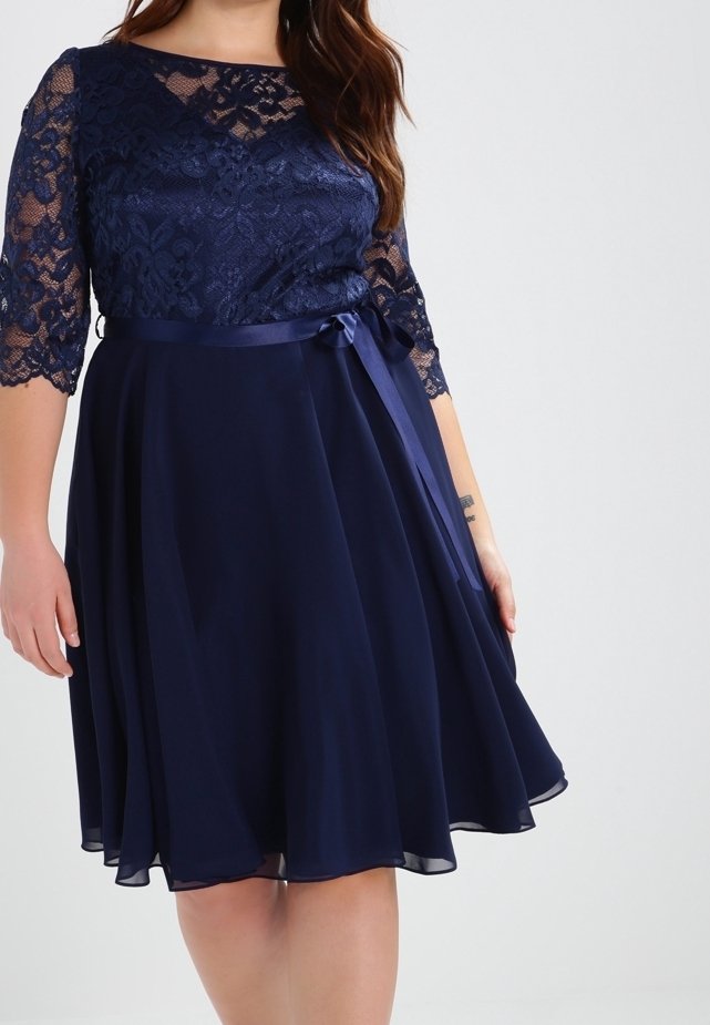 Robe bleu marine avec corsage et manches en dentelle, ruban de satin à la taille noué sur le côté, et jupe fluide mi-longue portée par une personne.