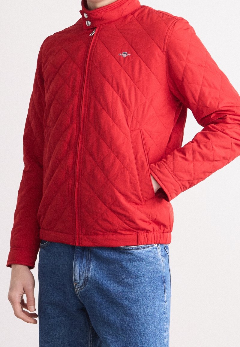 Giacca trapuntata rossa con colletto alto, chiusura con zip e due tasche laterali. Presenta un logo discreto sul petto e polsini a costine.