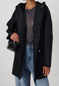Černá nepromokavá bunda se zipem, nastavitelným šňůrkovým pasem a kapucí. V kombinaci s průsvitným topem a modrými džíny s vysokým pasem.