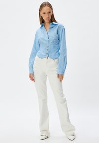 Camicia button-up a righe blu chiaro con maniche lunghe e design cropped, abbinata a pantaloni bianchi a vita alta e scarpe bianche a punta.