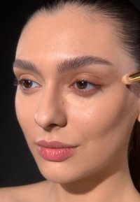 Model with smooth, even skin tone and natural makeup applying a light shade near the eye with a brush. Dark background enhances details.