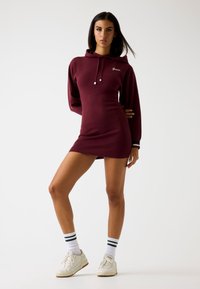 Abito felpa bordeaux con maniche lunghe, polsini a costine e logo frontale. Indossato con sneakers bianche e calzini a righe, mette in mostra una silhouette aderente.