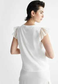T-shirt bianco con maniche a frill in rete, scollo rotondo e una superficie liscia. Il retro presenta un design semplice con un orlo leggermente curvo.