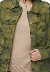 Camouflage-mønstret grøn jakke lavet af slidstærkt stof, med seks grønne knapper, en brystlomme og en afslappet pasform over en beige strikket sweater.