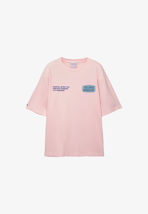 Vaaleanpunainen puuvillapaita, jossa on suuri mitoitus, lyhyet hihat ja sininen painettu teksti. Edessä on suorakulmainen logomuotoilu.