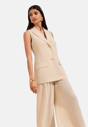 Femeie purtând un blazer bej fără mâneci, cu nasturi aurii, și pantaloni largi asortați, stilizate cu cercei și brățară aurii.