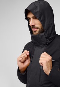 Zwarte waterdichte jas met een capuchon, heeft een hoge kraag, elastische trekkoorden en ritszakken aan de voorkant. Glad textuur.