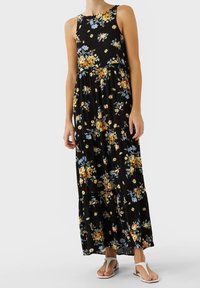 Vestido maxi negro con flores amarillas y azules, diseño sin mangas, falda en capas y tejido suave. Combinado con sandalias blancas.