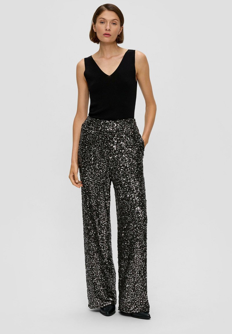 Schwarzes ärmelloses Oberteil kombiniert mit weiten, glitzernden Hosen in Schwarz und Silber, mit Taschen und hoher Taille. Schlichte schwarze Schuhe.