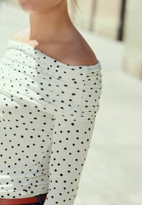 Top bianco con pois neri, design off-the-shoulder, texture arricciata, maniche lunghe, abbinato a una fascia rossa in vita.