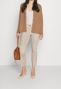 Beige cardigan over een lichte top, gecombineerd met lichte suèdebroek en nude hakken. Een bruin handtas met een kettinghandvat wordt in een hand vastgehouden.