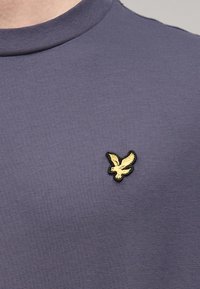 Μπλε ναυτικό κοντομάνικο t-shirt από λείο ύφασμα με στρογγυλή λαιμόκοψη, διακοσμημένο με κεντημένο χρυσό και μαύρο λογότυπο αετού στο στήθος.