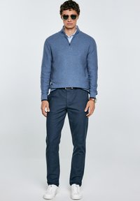 Suéter de cuello alto azul marino, camisa de cuello clara debajo, pantalones oscuros, zapatillas blancas y gafas de sol. Textura suave, diseño casual.