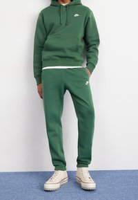 Person i grön Nike-huvtröja och matchande svettbyxor med beige höga sneakers står mot en vit vägg på en blå matta.