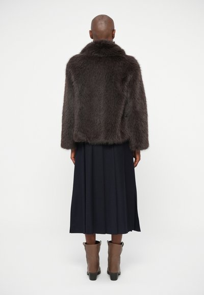 Casaco de pêlo sintético em castanho escuro com um colarinho alto, combinado com uma saia plissada azul marinho e botas de tornozelo castanhas. Aparência suave e texturizada.
