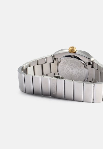 Reloj de metal plateado con una correa texturizada, que presenta una caja redonda y una corona dorada. La parte posterior muestra un emblema de Medusa y la inscripción de la marca.