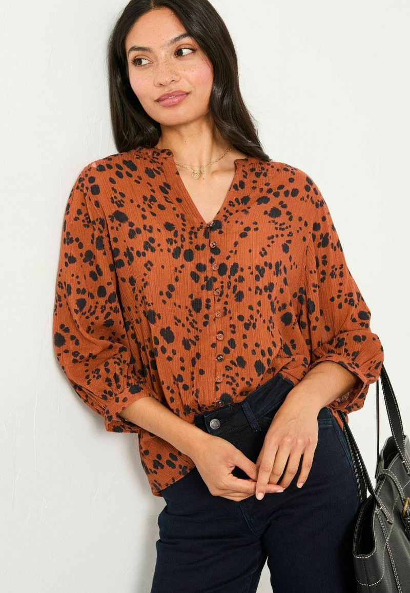 Rust-colored blouse with black spotted pattern, V-neck, button front, and loose sleeves, paired with dark jeans and a black handbag.