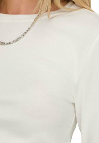 Weißes Langarmoberteil aus weichem Baumwollstoff, mit Rundhalsausschnitt und einer silbernen Kettenkette als Akzent. Leichte Textur sichtbar.