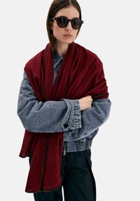 Sciarpa a quadri rossa avvolta intorno a una giacca di denim grigia. La sciarpa presenta un bordo sfrangiato, con una texture accogliente e un contrasto di colori caldi.