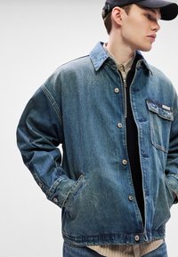 Τζάκετ από τζιν σε ξεθωριασμένο μπλε χρώμα με κουμπιά μπροστά, με�γάλη χάρτινη τσέπη και οξύαιχμο γιακά. Διαθέτει φαρδιά γραμμή και εμφανείς ραφές.