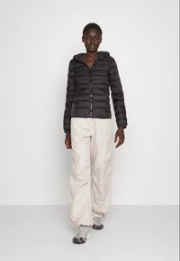 Casaco puffer preto com fecho e capuz, combinado com calças cargo em bege claro. Ténis cinzentos completam o conjunto, apresentando materiais texturizados.