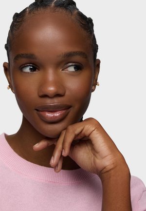 Mujer con el cabello trenzado, lleva pequeños pendientes de oro y un suéter rosa, apoyando la barbilla en la mano y mirando pensativamente hacia un lado.