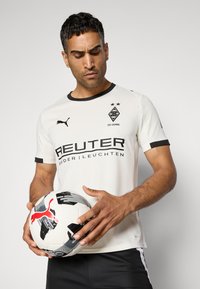 Hvid fodboldtrøje med sorte detaljer, der har et logo, tekst og rund halsudskæring. Spilleren holder en sort og hvid fodbold.