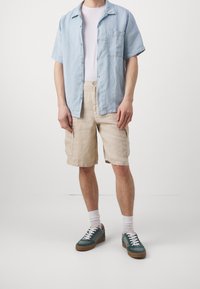 Camicia azzurra chiara a maniche corte con bottoni, t-shirt bianca sotto, pantaloni cargo beige e sneakers turchesi con lacci bianchi e suola marrone.
